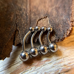 May include: Three silver curved barbell body jewelry pieces with ball ends. The jewelry is arranged in a row on a wooden surface.