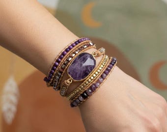 Puede incluir: Una pulsera envolvente de varias vueltas con una gran piedra de amatista morada de forma ovalada y facetada, engastada en metal dorado. La pulsera está hecha con cordón de cuero color canela natural, pequeñas cuentas moradas y detalles metálicos dorados.