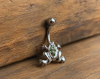 Puede incluir: Un piercing para el ombligo de tono plateado con un dije en forma de rana. La rana tiene un acabado metálico liso y está decorada con cuatro pequeñas piedras preciosas verdes circulares incrustadas en su espalda, con una barra curva.