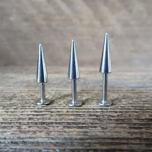 May include: Three silver-colored, cone-shaped body jewelry studs. Each stud has a pointed top and a flat, circular base. The studs are arranged in a row, with the middle one being the tallest. The background is a wooden surface.