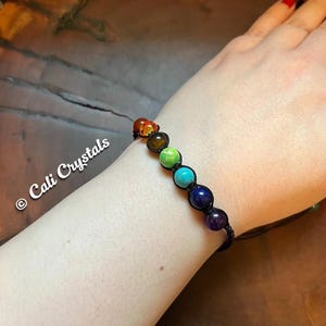 May include: A beaded bracelet featuring seven colorful gemstones, each representing a chakra. The beads are strung on a black cord. The bracelet is shown on a wrist. The text "Cali Crystals" is visible.