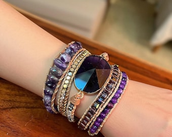 Puede incluir: Una pulsera envolvente de varias vueltas con cuentas de amatista moradas, detalles metálicos en tono dorado y una gran piedra central de color morado oscuro facetada. Las cuentas están sujetas con cordón de cuero beige claro.