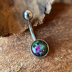 May include: A silver belly button ring with a round green, pink, and black gemstone.