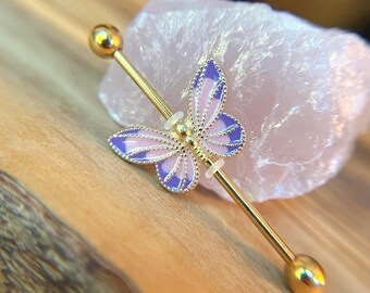 Puede incluir: Una joya corporal tipo barra industrial en tono dorado con un dije de mariposa con alas de esmalte morado y rosa claro. La mariposa está centrada en la barra metálica recta, que termina con una esfera lisa en cada uno de sus extremos.
