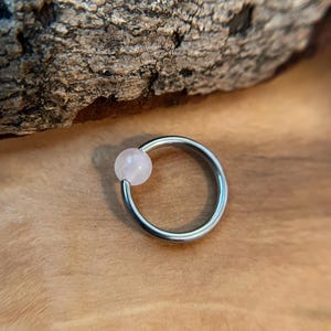 May include: A silver-colored captive bead ring with a light pink, round bead. The jewelry is set against a wooden surface with a piece of bark in the background. The ring is a type of body piercing jewelry.