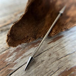 Puede incluir: Una joya de piercing corporal de tipo barbell industrial de color plateado. La barra recta est&aacute; rematada con un cono puntiagudo en cada extremo. La joya se muestra sobre una superficie de madera.