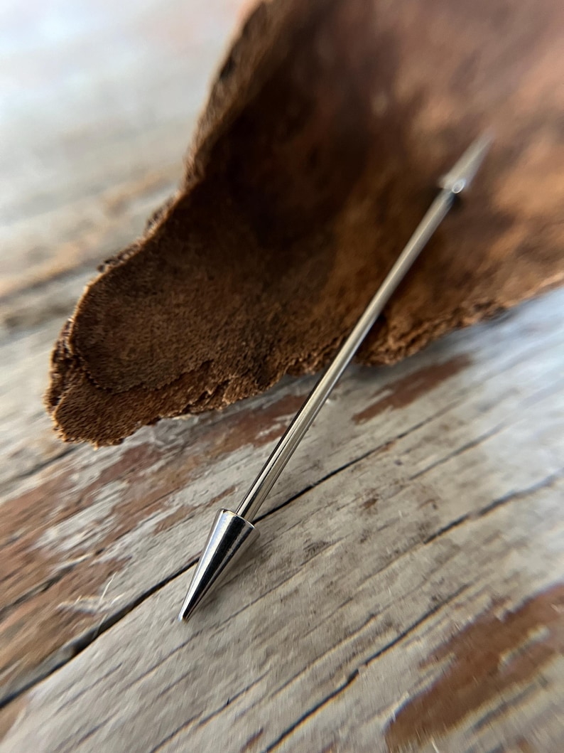 Puede incluir: Una joya de piercing corporal de tipo barbell industrial de color plateado. La barra recta est&aacute; rematada con un cono puntiagudo en cada extremo. La joya se muestra sobre una superficie de madera.