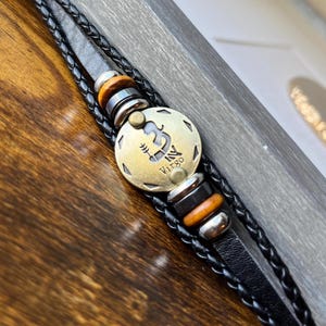 May include: Black leather bracelet with a gold-toned Virgo zodiac sign charm and brown and silver beads.