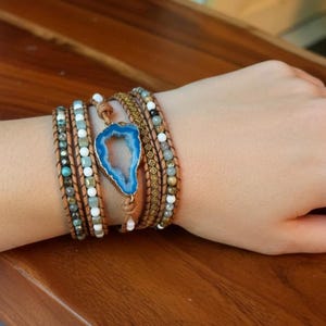 May include: A multi-strand wrap bracelet featuring brown leather cord, small faceted beads in shades of blue, white, and gray, and a central blue geode slice with a gold-toned edge. The bracelet is designed to wrap around the wrist multiple times.