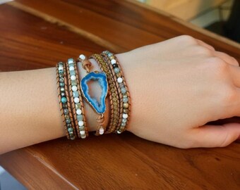 Puede incluir: Una pulsera envolvente de varias vueltas con cordón de cuero marrón, pequeñas cuentas facetadas en tonos azul, blanco y gris, y una rodaja de geoda azul central con un borde dorado. La pulsera está diseñada para rodear la muñeca varias veces.