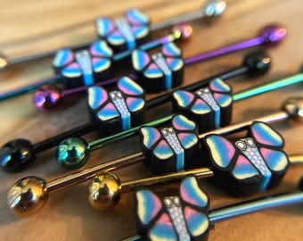 Puede incluir: Una colección de joyas corporales tipo barra industrial con dijes en forma de mariposa. Cada dije tiene una base negra con alas iridiscentes multicolores y un centro texturizado. Las barras vienen en varios acabados metálicos, incluyendo oro, plata, negro y tonos tornasolados.