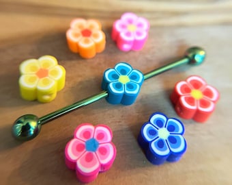 Puede incluir: Una joya corporal de barra industrial metálica con un acabado verde iridiscente, que presenta una cuenta extraíble en forma de flor azul en el eje, rodeada por seis cuentas sueltas en forma de flor en colores naranja, rosa, amarillo, rojo y morado.