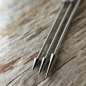 Puede incluir: Primer plano de cuatro pinchos de c&oacute;ctel plateados con puntas c&oacute;nicas. Los pinchos est&aacute;n dispuestos en diagonal sobre una superficie de madera clara y envejecida. Los pinchos met&aacute;licos tienen un acabado pulido y reflectante.