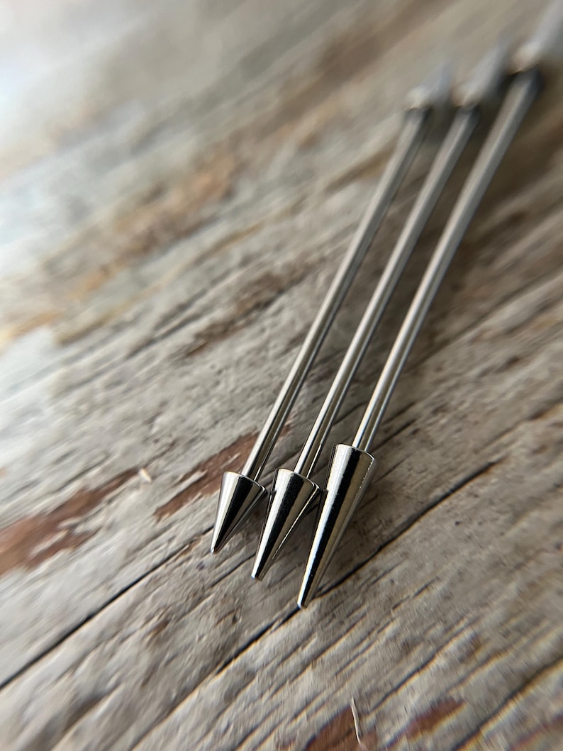 Puede incluir: Primer plano de cuatro pinchos de c&oacute;ctel plateados con puntas c&oacute;nicas. Los pinchos est&aacute;n dispuestos en diagonal sobre una superficie de madera clara y envejecida. Los pinchos met&aacute;licos tienen un acabado pulido y reflectante.