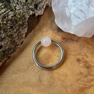 May include: A silver-colored captive bead ring with a light pink, round rose quartz bead. The ring is set against a wooden surface with a piece of bark and a chunk of rose quartz in the background.