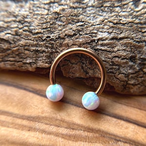 May include: A gold-colored horseshoe-shaped piercing ring with two iridescent, light blue and white opal-like beads. The jewelry is set against a textured wooden background.