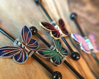 Puede incluir: Una colección de piercings industriales tipo barra con metal negro y extremos esféricos. Cada barra está adornada con un dije en forma de mariposa en varios colores de esmalte, incluyendo azul, morado, rojo, verde y rosa claro con bordes plateados.