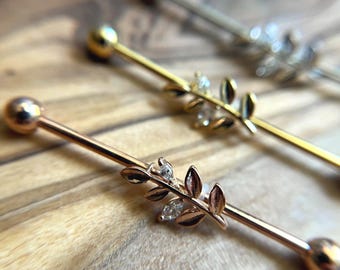 Puede incluir: Tres piercings industriales que consisten en una barra de metal recta con una bola en cada extremo. Cada barra está adornada con un adorno central en forma de hoja con pequeñas piedras transparentes. El conjunto incluye tonos oro rosa, oro y plata.