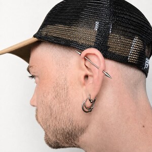 Puede incluir: Gorra de camionero de malla negra con visera marr&oacute;n. La persona lleva varios pendientes plateados, incluyendo aros y un piercing industrial. La persona tiene barba corta y la cabeza rapada.
