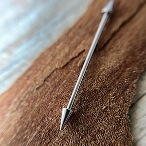 Puede incluir: Un piercing industrial plateado con extremos puntiagudos. La joya corporal se muestra sobre un fondo marr&oacute;n texturizado. La barra es recta con una punta c&oacute;nica en cada extremo.