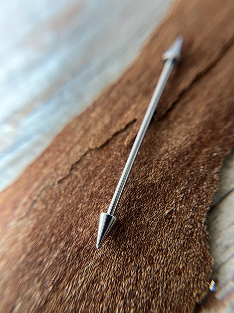 Puede incluir: Un piercing industrial plateado con extremos puntiagudos. La joya corporal se muestra sobre un fondo marr&oacute;n texturizado. La barra es recta con una punta c&oacute;nica en cada extremo.