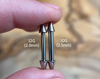 Puede incluir: Dos barras rectas de metal plateado con extremos en forma de cono puntiagudo. La barra de la izquierda está etiquetada como 12G (2,0 mm) y la de la derecha como 10G (2,5 mm). Estas piezas de joyería corporal tienen un acabado liso y pulido.