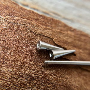 Puede incluir: Primer plano de tres piezas de joyer&iacute;a corporal de metal plateado. Dos piezas tienen forma c&oacute;nica con un extremo roscado, y la tercera es una barra recta con un extremo roscado. El fondo es una superficie marr&oacute;n texturizada.