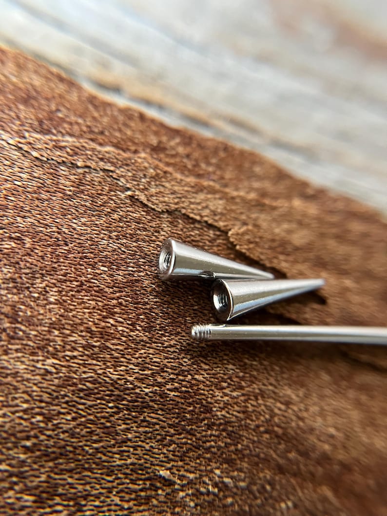 Puede incluir: Primer plano de tres piezas de joyer&iacute;a corporal de metal plateado. Dos piezas tienen forma c&oacute;nica con un extremo roscado, y la tercera es una barra recta con un extremo roscado. El fondo es una superficie marr&oacute;n texturizada.
