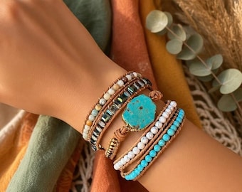 Puede incluir: Una pulsera envolvente de varias vueltas con una piedra turquesa central, hileras de cuentas blancas y azul brillante, detalles metálicos gris oscuro y pequeñas piedras en tonos tierra, unidas con cordón de cuero marrón.
