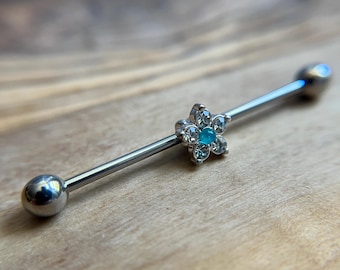 Puede incluir: Una joya corporal tipo barra industrial de tono plateado con un dije central en forma de flor. La flor tiene cinco pétalos de cristal transparente que rodean una pequeña piedra redonda de color turquesa brillante en el centro.