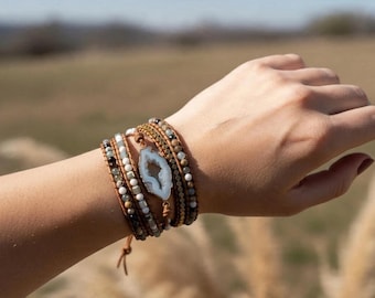 Puede incluir: Una pulsera envolvente de varias vueltas con una rodaja de geoda blanca central, rodeada de pequeñas cuentas redondas en tonos marrón, gris, blanco y oro metálico, ensartadas en un cordón de cuero marrón con cierre de botón.