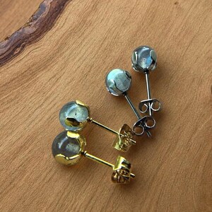 May include: Close-up of two pairs of stud earrings. One pair has gold posts and settings, the other has silver posts and settings. The earrings feature round, iridescent, light blue-gray stones. The earrings are displayed on a wooden surface.