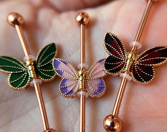 Puede incluir: Tres piercings de barra industrial con barras de metal en tono oro rosa y extremos esféricos. Cada barra está adornada con un dije de mariposa en esmalte verde, morado claro o rojo oscuro, con detalles y bordes en metal dorado.