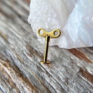May include: A small, gold-colored wind-up key. The key has a T-shaped design with a circular base and two eyelets at the top. It is set against a backdrop of a light-colored stone and a weathered wooden surface.