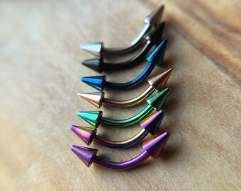Puede incluir: Una colección de seis piezas de joyería corporal tipo barra curva dispuestas en una fila vertical. Cada pieza presenta un eje de metal curvo y liso con una punta cónica afilada en ambos extremos. Los colores incluyen plata, negro, azul, oro, verde y morado.