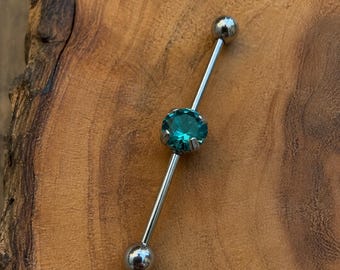 Puede incluir: Una pieza de joyería corporal tipo barra industrial plateada con una piedra preciosa redonda y facetada de color verde azulado engastada en el centro. La barra tiene un eje de metal liso con una bola esférica pulida en cada extremo.