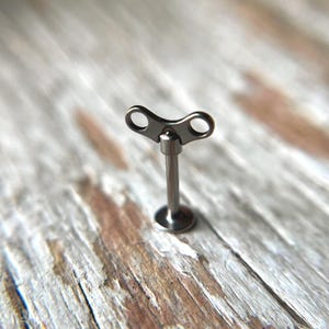 May include: A small, silver-colored wind-up key, featuring a circular base, a slender stem, and a top with two eye-shaped openings. The key is set against a blurred, light-colored wooden surface.