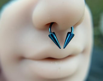 Puede incluir: Un aro de septum azul metálico con un diseño circular y dos puntas cónicas en los extremos. La joya está colocada en el centro de la nariz, mostrando un acabado liso y pulido junto con un color azul intenso y vibrante.