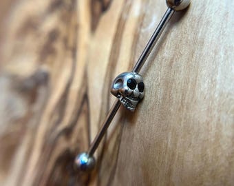 Puede incluir: Una joya corporal tipo barra industrial de tono plateado con un dije central de calavera con ojos negros incrustados. La barra tiene un acabado metálico liso e incluye dos tapas esféricas que se enroscan en el extremo de la pieza.