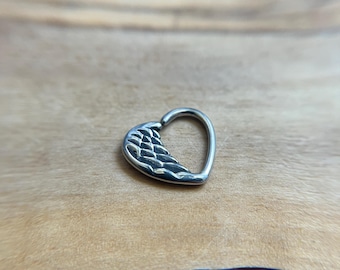 Puede incluir: Un pendiente de aro de metal plateado con forma de corazón, que presenta un diseño de ala texturizada en un lado. La pieza de joyería tiene un acabado liso y pulido en el lado liso del corazón y un patrón de plumas grabado detallado en el ala.