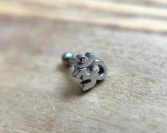 Puede incluir: Una joya corporal de metal plateado con un diseño de símbolo Om en tres dimensiones. La pieza incluye un pequeño cierre esférico unido a un poste recto, con el emblema Om sirviendo como el elemento decorativo frontal de la pieza.