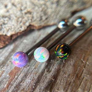 May include: Close-up of three industrial barbells with colorful opal-like ball ends. The balls are purple, white, and black with iridescent patterns. The barbells are silver and rest on a textured wooden surface.
