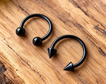 May include: Two black circular barbell body jewelry pieces. One piece features two smooth, round ball ends, while the other piece features two sharp, conical spike ends. Both items are constructed from a solid black metal material with a glossy finish.