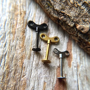 May include: Three small, decorative wind-up keys in black, gold, and silver, arranged on a weathered wooden surface. The keys have a classic design with a circular base and a keyhole-shaped top. A piece of wood is in the background.