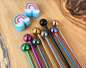 Puede incluir: Una colección de barras de piercing de titanio IP en varios acabados metálicos incluyendo oro, plata, negro, oro rosa y arcoíris iridiscente, acompañadas de dos dijes azul claro en forma de arcoíris con el texto Pick Your Color Titanium IP Barbell.