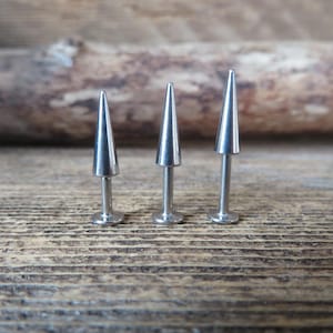May include: Three silver-colored, cone-shaped lip piercing studs. The studs have a flat base and a pointed top. The background is a wooden surface.