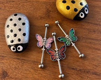 Puede incluir: Tres piercings industriales con dijes de mariposa en esmalte rosa, rojo y verde, acompañados de dos piedras pintadas como mariquitas, una plateada con puntos negros y otra dorada con puntos negros, dispuestos sobre una superficie de madera.