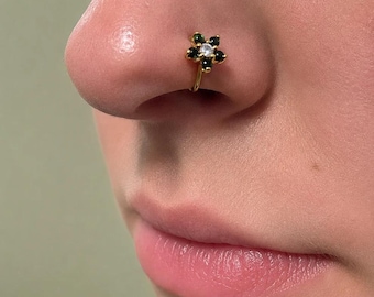 Puede incluir: Un aro para la nariz de tono dorado con un diseño en forma de flor. El centro de la flor es una piedra redonda transparente, rodeada por cinco piedras redondas de color verde oscuro que forman los pétalos. La joya está colocada en la aleta nasal.