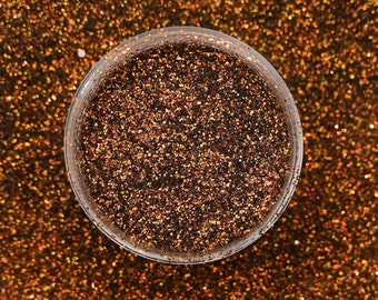 May include: A close-up of a small jar filled with fine, sparkly glitter. The glitter is a mix of brown, orange, and gold colors.