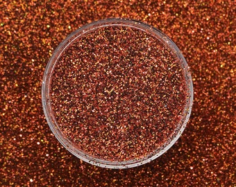 May include: A clear, round container filled with copper-colored glitter. The glitter is scattered across the surface, creating a sparkling effect. The glitter is a mix of different shades of copper and bronze.